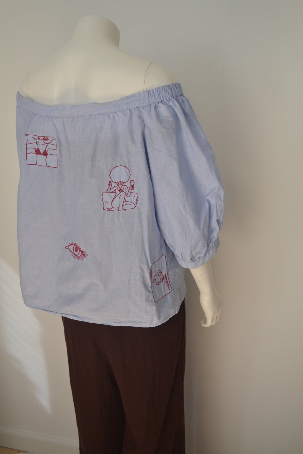 Lys blå off-shoulder bluse bagfra med bordeauxrøde broderede motiver — bikini, dame med afro, øje og spillekort